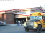 Foto do exterior de uma escola de ensino fundamental. Veem-se crianas chegando e um nibus de transporte de estudantes.  Palavras-chave: colgio, crianas, primrio.