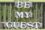 Nesta imagem, v-se a foto de uma placa com a expresso "Be my guest" (Fique  vontade), em frente a uma cerca de madeira.   Palavras-chave: Recepo. Cortesia. Idioms. Expresso idiomtica.