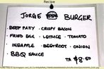 Foto de um cartaz com os ingredientes de um sanduche (hambrguer, bacon, ovo frito, alface, tomate, abacaxi, beterraba, cebola e molho barbecue) e o respectivo preo.  Palavras-chave: Junk food. Alimentao. Recipe. Numerais.