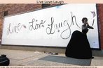 Foto de um outdoor preso a uma parede, tendo ao lado uma manequim. No outdoor se leem as palavras: "Live, Love, Laugh", nome de uma msica do cantor Clay Walker. A sequncia tambm se encontra num jingle (dos anos 1950) da rede de restaurantes estadunidense, som esse disponibilizado na pgina da TV Multimdia. Palavras-chave: Country. Cidade. Grafite. Lyrics. 