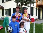Foto de uma família com quatro filhos, no exterior de uma casa, usando vários objetos esportivos.  Palavras-chave: expressão facial, futebol, faixas, esportes, Alemanha, Itália, casal, choro, criança, cores. 