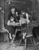 Figura de três crianças próximas a uma mesa, à qual um idoso está sentado. A menina lê, e o homem fuma seu cachimbo.  Palavras-chave: criança, mesa, idoso, leitura, cachimbo, família. 