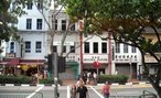 Foto de vários casarios coloniais em estilo espanhol, no centro de Singapura. Cidadãos cruzam uma rua. Em uma das fachadas, vê-se também uma placa com o nome "Rocha House".  Palavras-chave: cidade, trânsito, pedestres, colonização, interculturalidade.