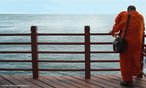 Foto de um monge budista recostado à amurada da praia de Sentosa, em Singapura. O homem permanece de costas para a câmera, que retrata ao fundo o Oceano Índico.  Palavras-chave: religioso, mar, natureza, paisagem, céu, solidão, tranquilidade.