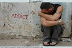 Foto de um rapaz agachado junto ao muro onde se lê a palavra "addict" (viciado). Palavras-chave: Drogadito. Vício. Drogas. Jovem. Efeito.