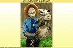 Foto antiga (colorizada) de um homem de idade, sorridente, segurando um possum, tipo de marsupial endêmicos da Austrália. Palavras-chave: Marsupial. Animais. Possum. Campo semântico. Austrália. Nova Guiné. Celebes. Descrição.