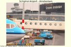 Foto da área de embarque de cargas e passageiros, no aeroporto de Schiphol, em Amsterdã. Vê-se, além da torre de controle, parte de uma aeronave e vários veículos de transporte de malas. Palavras-chave: Trabalho. Viagem. Estrutura. Holanda. Meios de transporte.