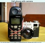 Foto humorística de uma máquina fotográfica amarrada a um telefone celular de modelo antigo. Palavras-chave: Indústria. Comércio. Consumo. Tecnologia. Modernidade.